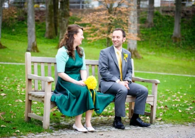 Brautpaar sitzt fröhlich auf Bank im Kurpark Bad Tölz, Hochzeitsfoto in natürlicher Umgebung.