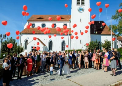 Hochzeit in Oberbayern: Brautpaar und Gaeste lassen rote Luftballons vor einer Kirche steigen. Hochzeitsfotografie.