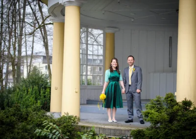 Brautpaar bei Hochzeit im Kurhaus Bad Tölz, posiert glücklich vor Pavillon mit gelben Säulen und Blumenstrauss.