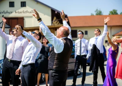 Gäste bei Hochzeit in Neukeferloh, heben Arme, lachen, Sommer, Hochzeitsfotografie im Freien vor Markthalle.