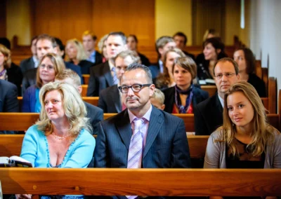Menschen in formeller Kleidung sitzen in Kirchenbänken; der Fokus liegt auf drei Personen in der vorderen Reihe.