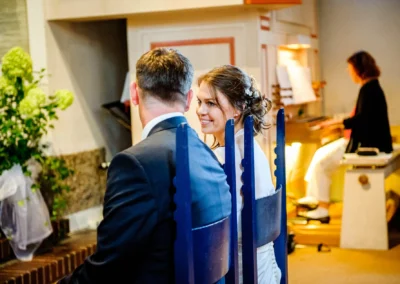 Brautpaar sitzt in Kirche, Braut lächelt seitlich zu Bräutigam, Organistin spielt im Hintergrund; grüne Blumen links.