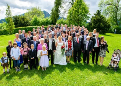 Hochzeitsgesellschaft im Freien beim Tegernsee: Gruppenfoto mit Brautpaar und Gaesten vor gruenem Hintergrund. Hochzeitsreportage.