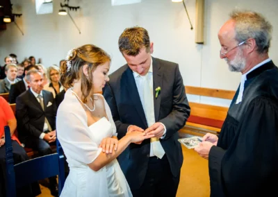 Ein Bräutigam steckt der Braut in einer Kirche den Ring an den Finger, während ein Geistlicher zusieht; Gäste sitzen gespannt.