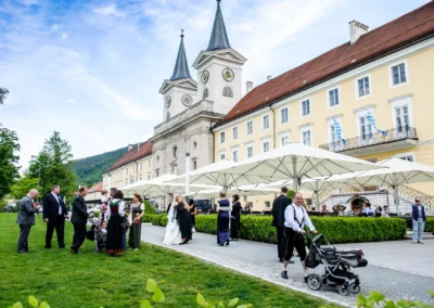 Hochzeit am Tegernsee: Gaeste im Garten einer Kirche, Hochzeitsfotografie in Oberbayern unter Sonnenschirmen.