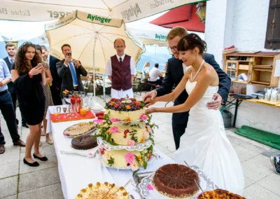 Brautpaar schneidet Hochzeitstorte bei Feier am Schliersee, Gaeste fotografieren den Moment, Hochzeitsfotografie.