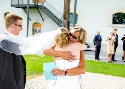 Braut umarmt Gast bei Hochzeit in Kloster Fuerstenfeld, Hochzeitsfotografie im Brucker Land, Gaeste im Hintergrund.