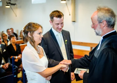 Brautpaar im formellen Outfit tauscht Ringe vor einem Geistlichen in einer Kirche, Gäste schauen zu, helle Beleuchtung.