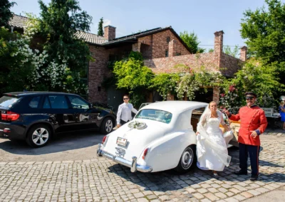 Braut steigt aus Hochzeitsauto vor Alter Gaertnerei Taufkirchen. Hochzeitsfoto mit Chauffeur und darueber rosengezierter Wand.