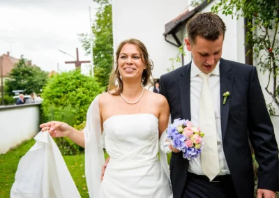 Brautpaar im Freien; sie trägt ein weißes Kleid mit Perlenkette, er einen dunklen Anzug; beide wirken fröhlich.