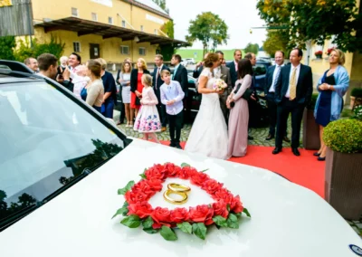 Hochzeitsfeier mit Brautpaar und Gästen vor Gasthof Stangl in Neufarn bei München. Hochzeit mit Blumenschmuck auf Auto.