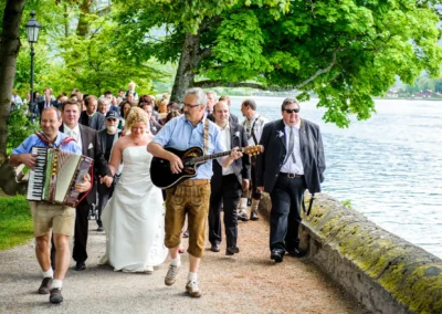 Hochzeitsgesellschaft mit Akkordeon und Gitarre spaziert am Tegernsee entlang, aufgenommen bei einer Hochzeit.