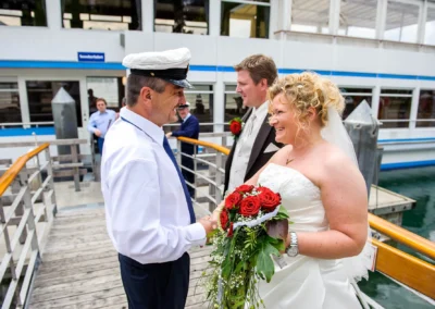 Brautpaar auf einem Steg am Tegernsee, lachend mit Kapitaen, Brautstrauss mit roten Rosen. Hochzeitsfotograf Bayern.