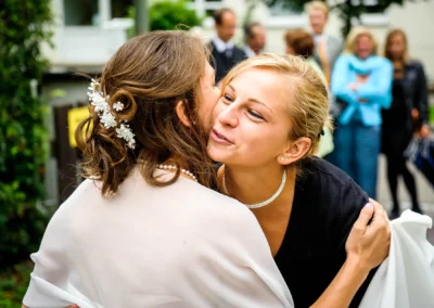 Zwei Frauen umarmen sich herzlich, die eine trägt eine weiße Bluse mit Perlenkette, die andere dunkle Kleidung und Schmuck. Im Hintergrund unscharf Menschen.