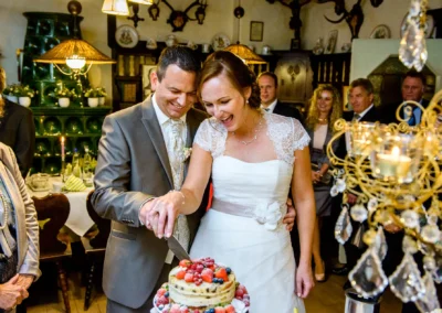 Brautpaar schneidet Hochzeitstorte im Gasthof Stangl an, mit Gästen im Hintergrund. Hochzeitsfotogene Momente.