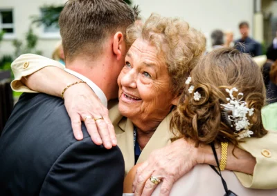 Ältere Frau umarmt zwei Personen, lächelt herzlich, trägt goldene Ringe und Armband; Hintergrund verschwommen und grün.