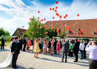 Hochzeit auf Gut Schloss Sulzemoos, Gäste lassen herzförmige Luftballons steigen, Brautpaar beobachtet.