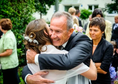 Mann im Anzug umarmt eine Frau, trägt Haaraccessoire; lächelnd, umgeben von Gästen im Freien, Hintergrund mit Grünpflanzen.