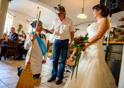 Braut in weissem Kleid lacht bei Hochzeit in Gasthof Widmann. Ein Mann kniet mit Besen, ein anderer spricht ins Mikrofon.