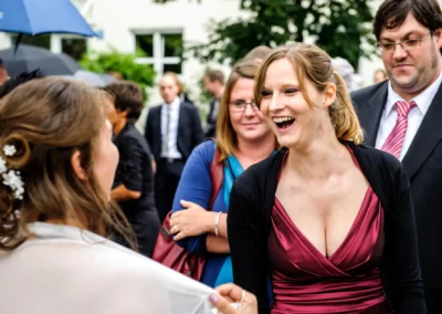 Lachende Frau in weinrotem Kleid, schwarzer Jacke, umgeben von Gästen im Freien, eine hat eine weiße Haarblume.
