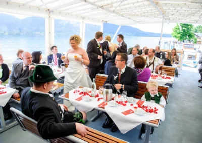 Hochzeitsfeier auf einem Boot am Tegernsee, Braut im Gespräch mit Gästen, stimmungsvoller Hochzeitsreportage-Moment.