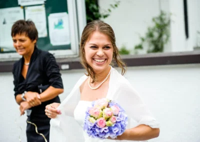 Lächelnde Frau im weißen Kleid mit Brautstrauß, daneben Person in schwarzer Kleidung, unscharfer Hintergrund mit grünen Pflanzen.