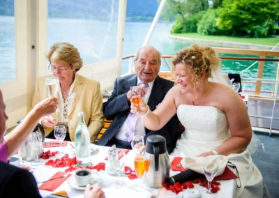 Braut mit Gaesten beim Hochzeitsessen auf dem Tegernsee, freudige Stimmung und Natur im Hintergrund.