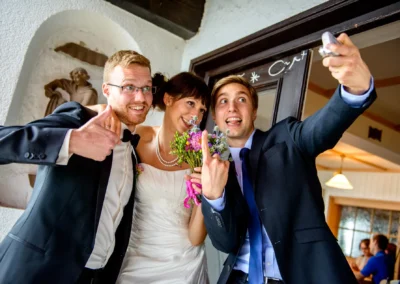 Brautpaar bei der Hochzeitsreportage im Kreis Miesbach, lacht und posiert fuer ein Selfie mit einem Freund.