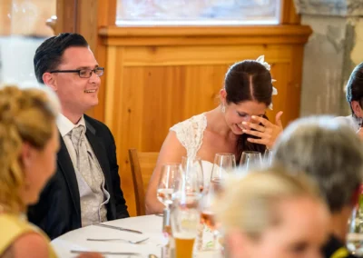 Hochzeitsfoto bei Gut Schloss Sulzemoos: Paar lacht am Tisch, umgeben von Gaesten und Weinglaesern.