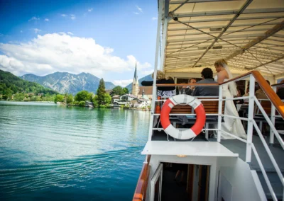 Hochzeitsreportage auf einem Boot am Tegernsee, Brautpaar mit Gästen, Berge im Hintergrund, Hochzeitsfotografie in Bayern.