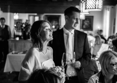 Brautpaar in festlicher Kleidung mit Sektgläsern; leuchtende Stimmung in gemütlichem Raum, Gäste im Hintergrund.