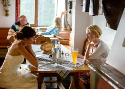 Braut in Kleid beim Gespraech mit Freundin in rustikaler Gaststube, Hochzeit am Schliersberg-Alm fotografiert.