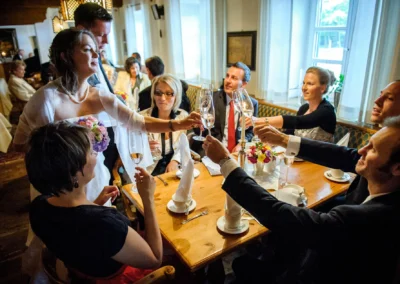 Braut erhebt Sektglas zum Anstoßen, umgeben von elegant gekleideten Gästen, stimmungsvolle Beleuchtung im Innenraum, Blumenstrauß in der Hand.