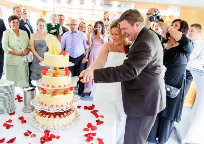 Brautpaar schneidet Hochzeitstorte bei Feier in Oberbayern, umgeben von Familie und Freunden. Hochzeit Tegernsee.