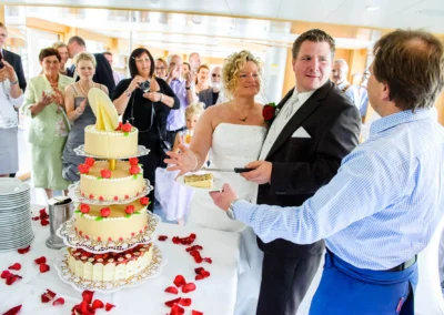 Brautpaar im eleganten Hochzeitskleid schneidet mehrstoeckige Torte an. Hochzeit in Tegernsee, Bayern.
