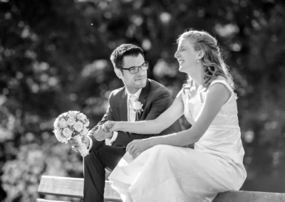 Brautpaar lacht auf Bank mit Blumenstrauss bei Hochzeit in Assenhausen, festgehalten in dieser Hochzeitsfotografie.