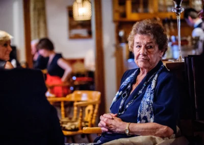 Ältere Frau in dunkler Kleidung sitzt in einem gemütlich beleuchteten Raum, umgeben von anderen Gästen.