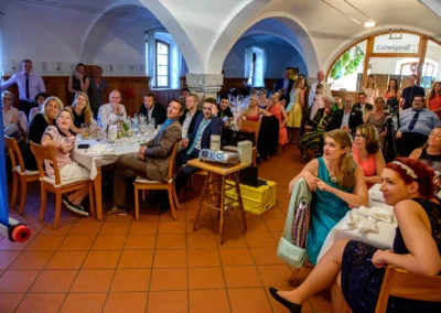 Gäste sitzen bei einer Hochzeit auf Gut Schloss Sulzemoos und schauen gespannt auf eine Präsentation. Hochzeitsfotografie.