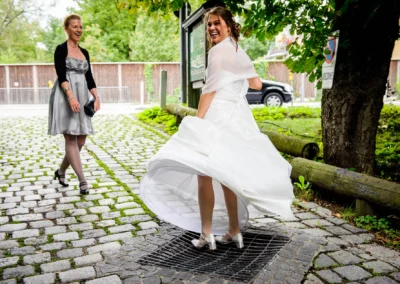 Braut in wirbelndem weißen Kleid auf Kopfsteinpflaster, lacht; Begleiterin in grauem Kleid und schwarzem Jäckchen lacht mit.
