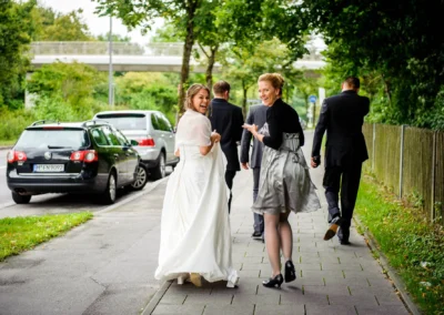 Braut im weißen Kleid und Frau im grauen Kleid lächeln auf Gehweg, begleitet von zwei Männern in Anzügen, parkende Autos im Hintergrund.