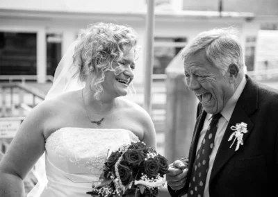 Braut mit Blumenstrauss lacht mit Gast bei Hochzeitsreportage in Bayern. Hochzeitsfotograf fängt Moment ein.