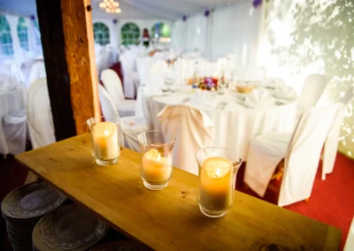 Kerzen auf einem Holztisch im Vordergrund, im Hintergrund elegant gedeckte Tische in einem festlich dekorierten Zelt.