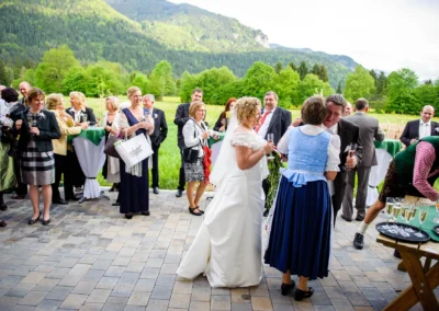 Hochzeitsfoto in Kreuth: Gäste im Freien, lachend bei Sektgläsern, Berge im Hintergrund, festliche Kleidung.