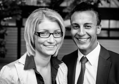 Zwei lächelnde Personen, eine mit Brille, im Freien, hinter ihnen verschwommene Bäume und ein Gebäude.