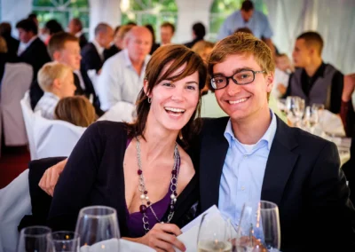 Lachendes Paar sitzt bei einer Feier an einem festlich gedeckten Tisch, umgeben von weiteren Gästen in eleganter Kleidung.