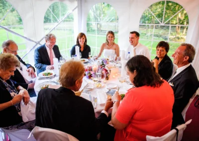 Acht Personen sitzen lächelnd an einem gedeckten Tisch in einem Festzelt, umgeben von grünem Gartenblick.