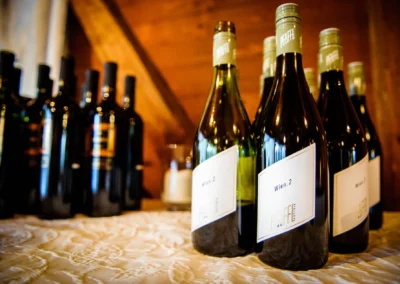 Mehrere Weinflaschen stehen auf einem bestickten Tuch, im Hintergrund unscharfe Flaschen auf Holzregal.