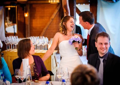 Eine Braut in weißem Kleid lacht mit einem Mann im Anzug; sie hält einen lila Blumenstrauß, umgeben von Gästen.