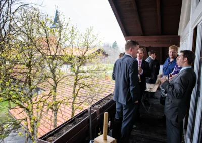 Gaste in Anzug auf Balkon bei einer Hochzeitsfeier in Hechenberg, fruhlingshafte Baume im Hintergrund. Hochzeitsfotografie.