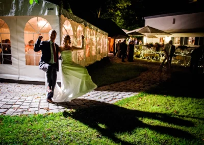 Paar tanzt freudig im Scheinwerferlicht vor beleuchtetem Zelt, wirft lange Schatten; umgeben von Partylichtern und Gästen.